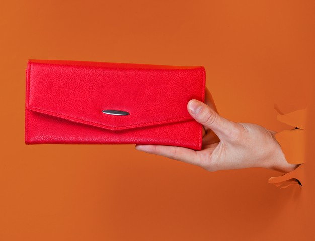 female-hand-holding-red-wallet-through-torn-orange-paper-minimalistic-creative-fashion-concept_175682-13337